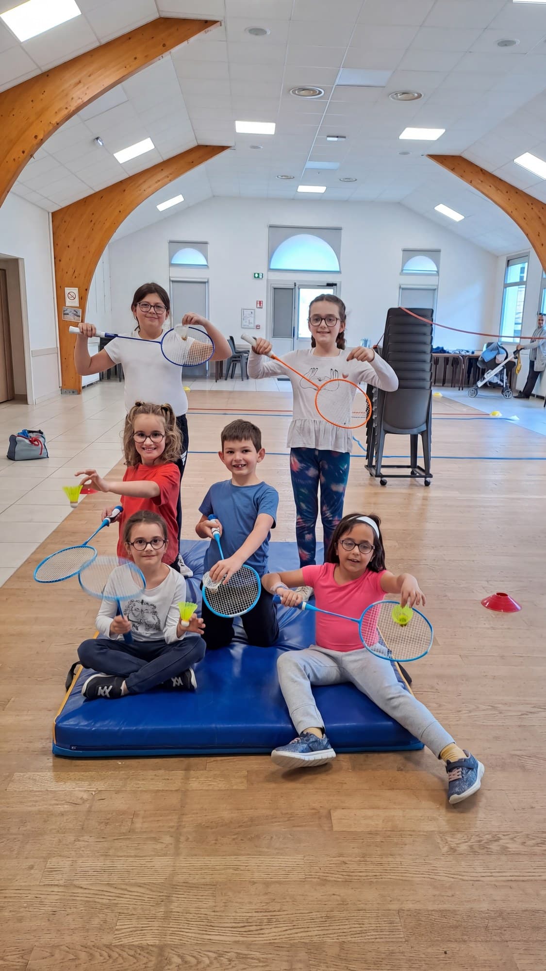Six enfants posent avec leurs raquettes de badminton, assis sur un tapis bleu.