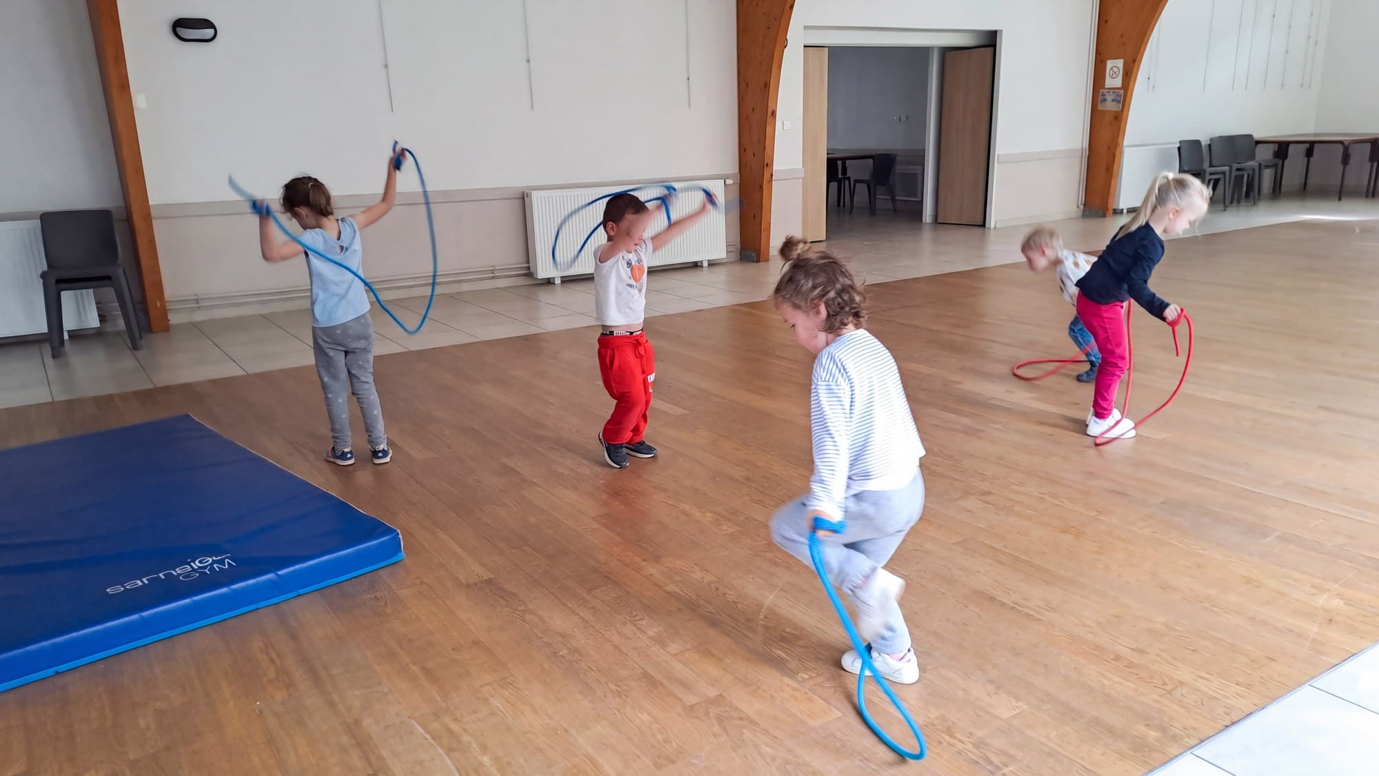Quatre enfants sautent à la corde dans la salle, un tapis bleu au sol.