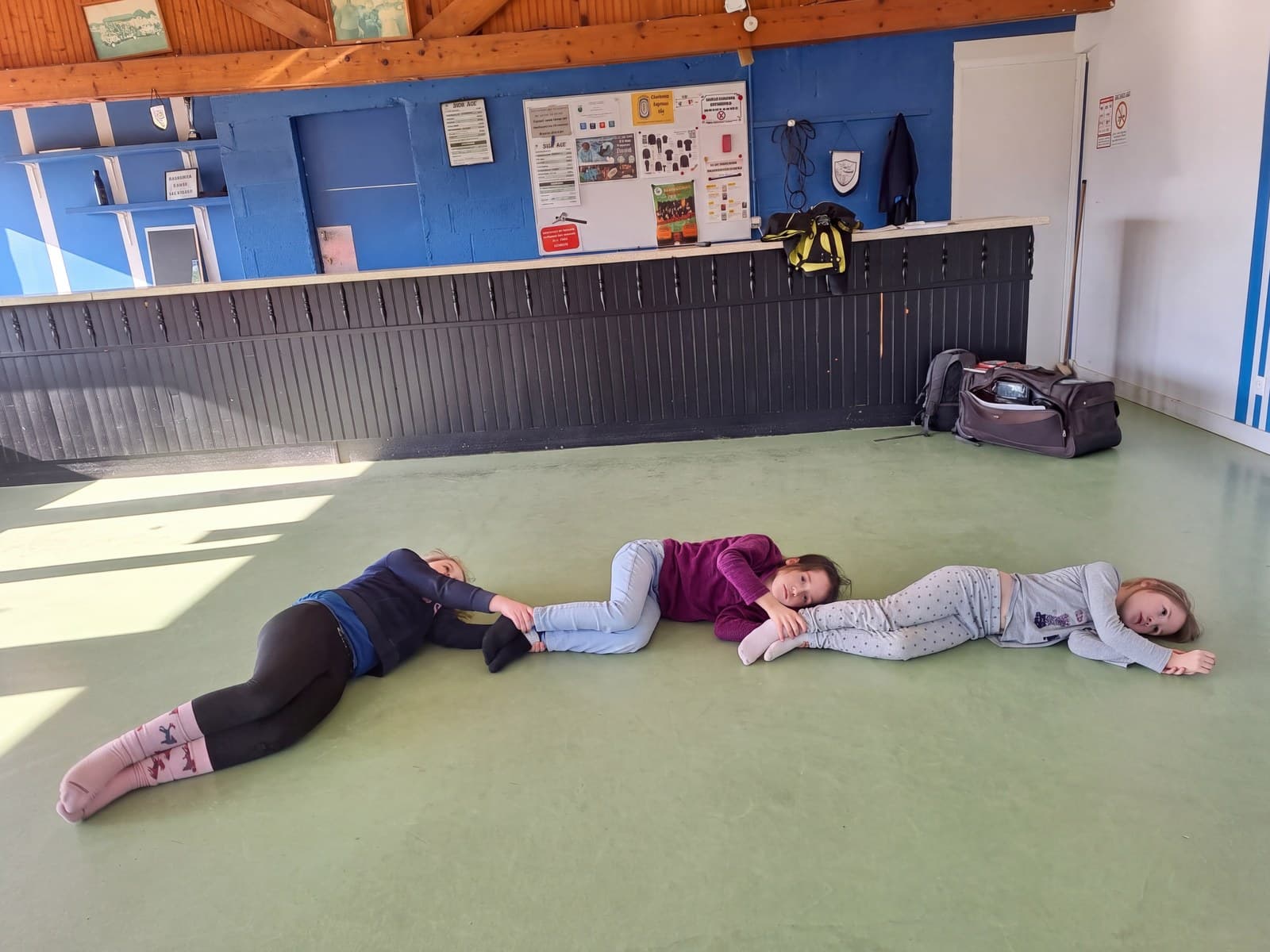 Trois filles allongées en ligne sur le sol, en étirement, dans une salle au sol vert.