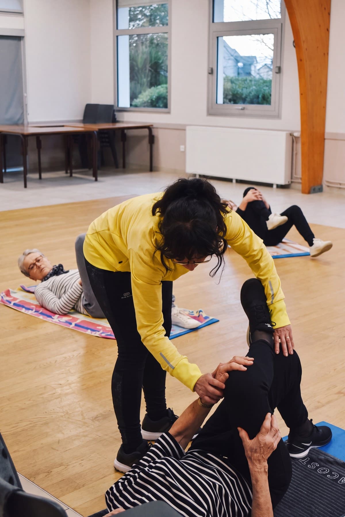 L’éducatrice aide une adhérente allongée sur son tapis, les autres travaillent derrière.