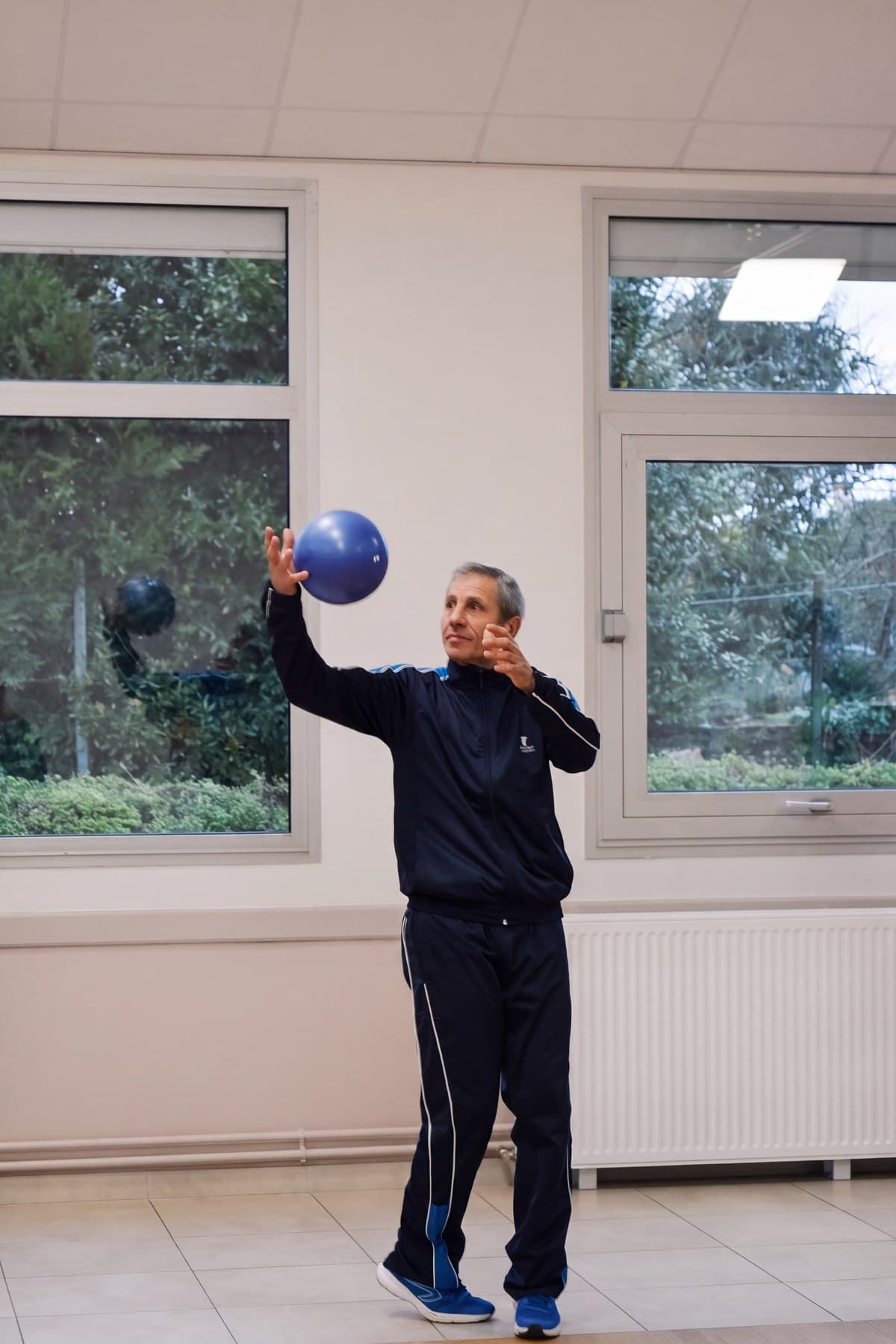 Un adhérent sénior lance un ballon bleu d’un bras, devant la fenêtre de la salle.